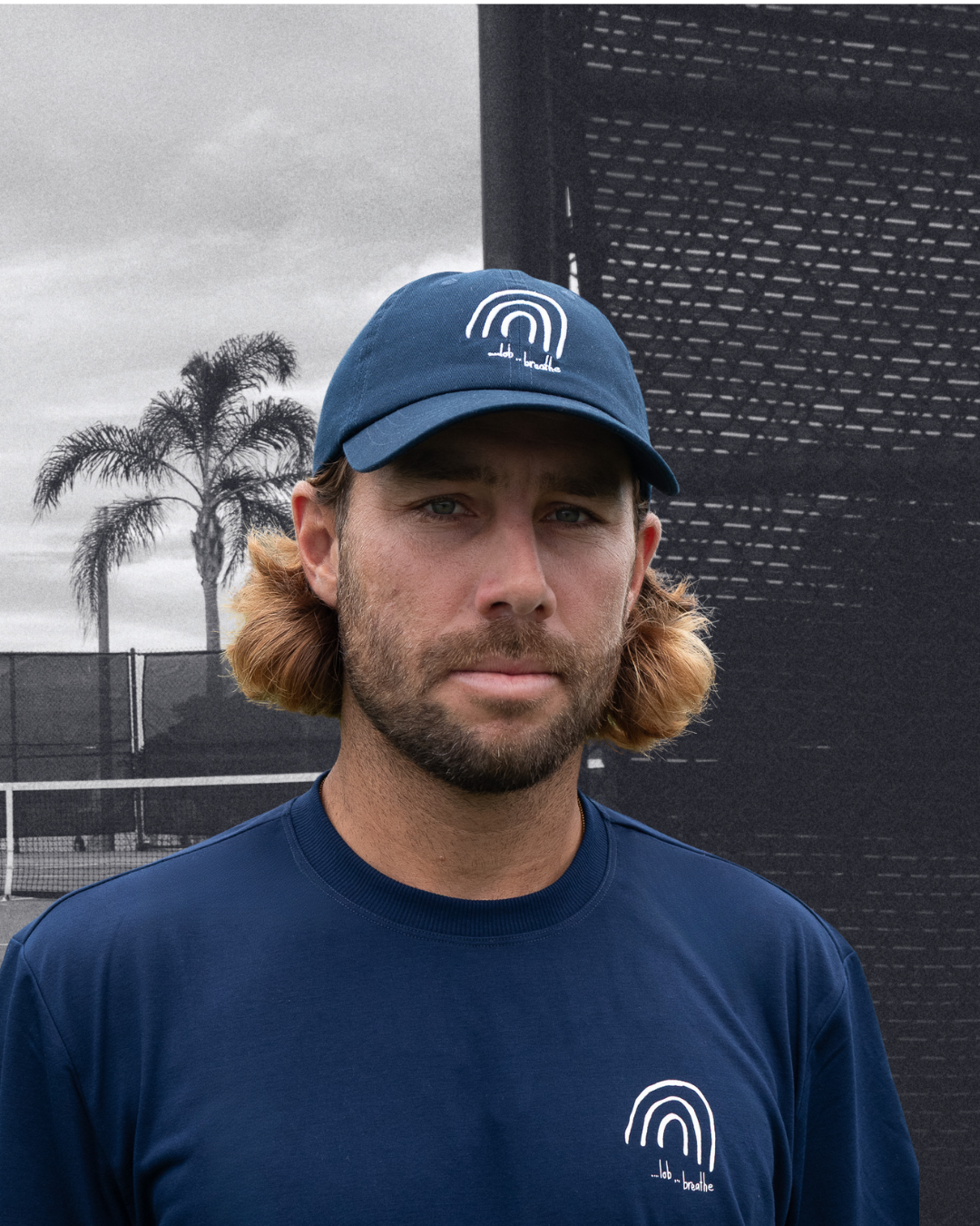 Man wearing a navy cap and t-shirt with a logo against a tennis court background