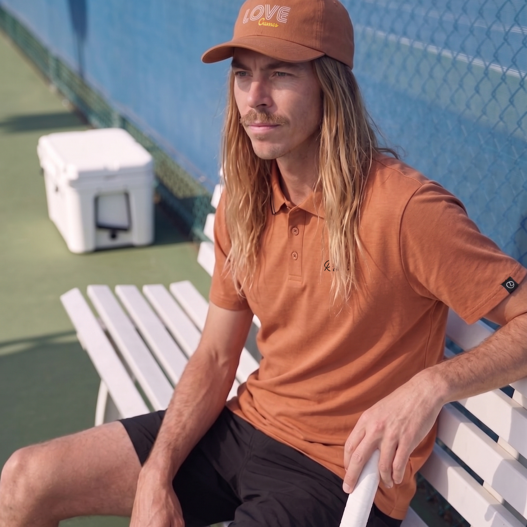 Man in clay polo and cap sitting on a tennis court with a racket.