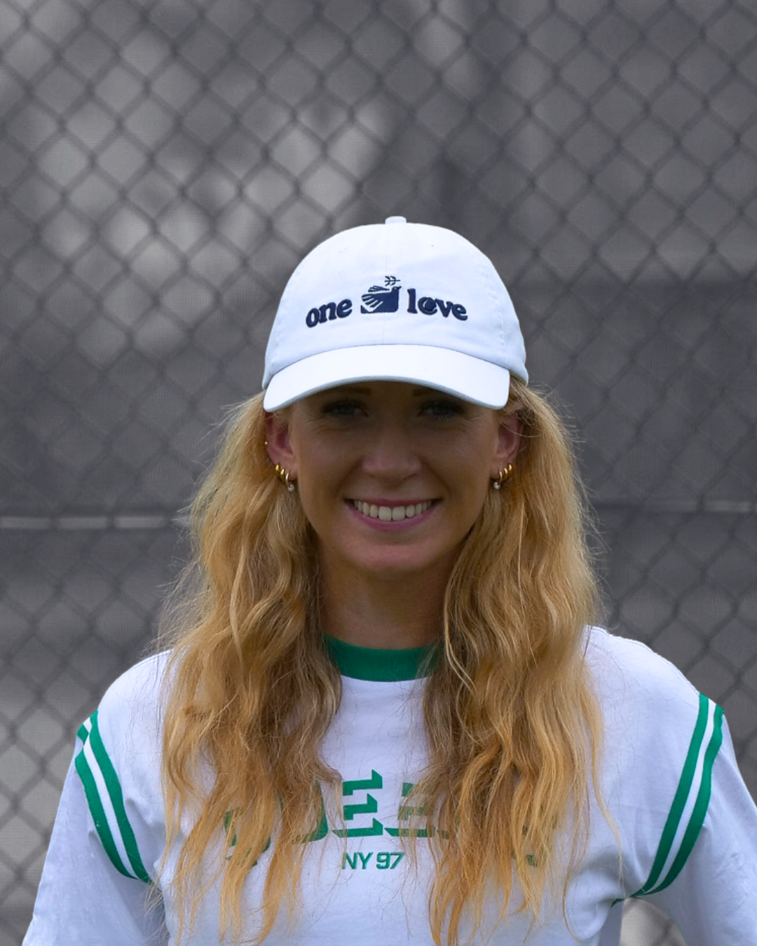 Person wearing a white cap with 'one love' text and a white shirt with green accents, standing in front of a tennis court