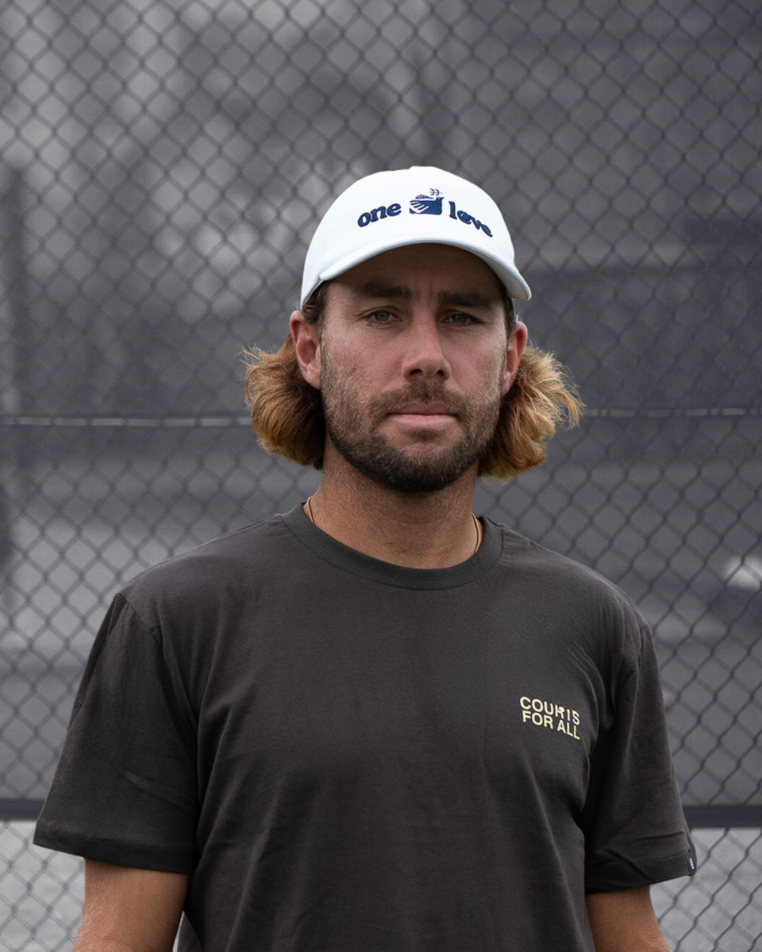 Man wearing a white cap and black t-shirt with text, standing in front of a tennis court