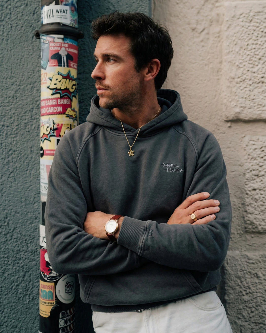 Man wearing a Rallee Quiet Please hoodie , standing against a textured wall with posters.