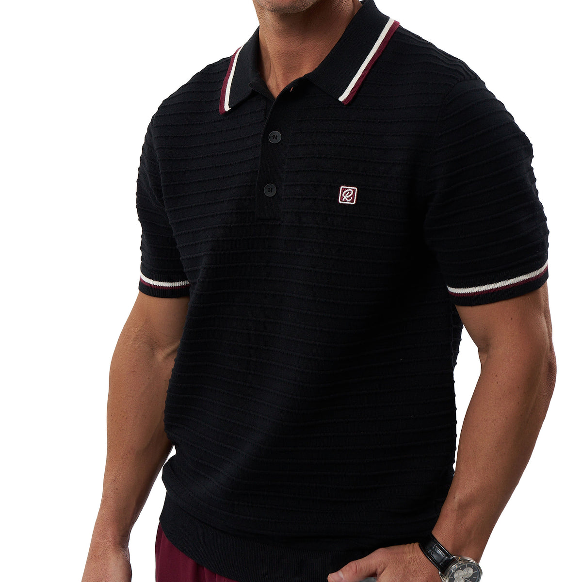 A man wearing a black short-sleeve polo shirt with a white and red striped collar and sleeve cuffs, featuring a small red square logo on the left chest.