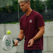 A man in a maroon short-sleeve athletic t-shirt with a small crest logo on the chest, holding a padel racket and preparing to hit a yellow tennis ball. The background shows a blurred outdoor court setting.