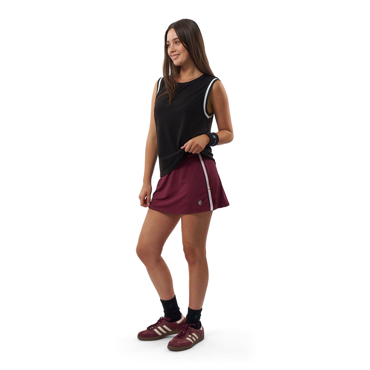 A woman modeling an athletic outfit consisting of a black sleeveless top with white trim and a maroon athletic skirt with a white side stripe, paired with maroon sneakers and black socks, against a plain white background.
