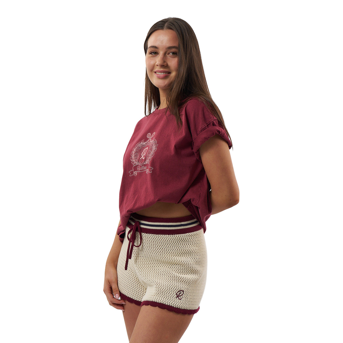 A woman modeling a maroon short-sleeve crop top with a white crest graphic and matching maroon-and-white knit shorts against a plain white background.
