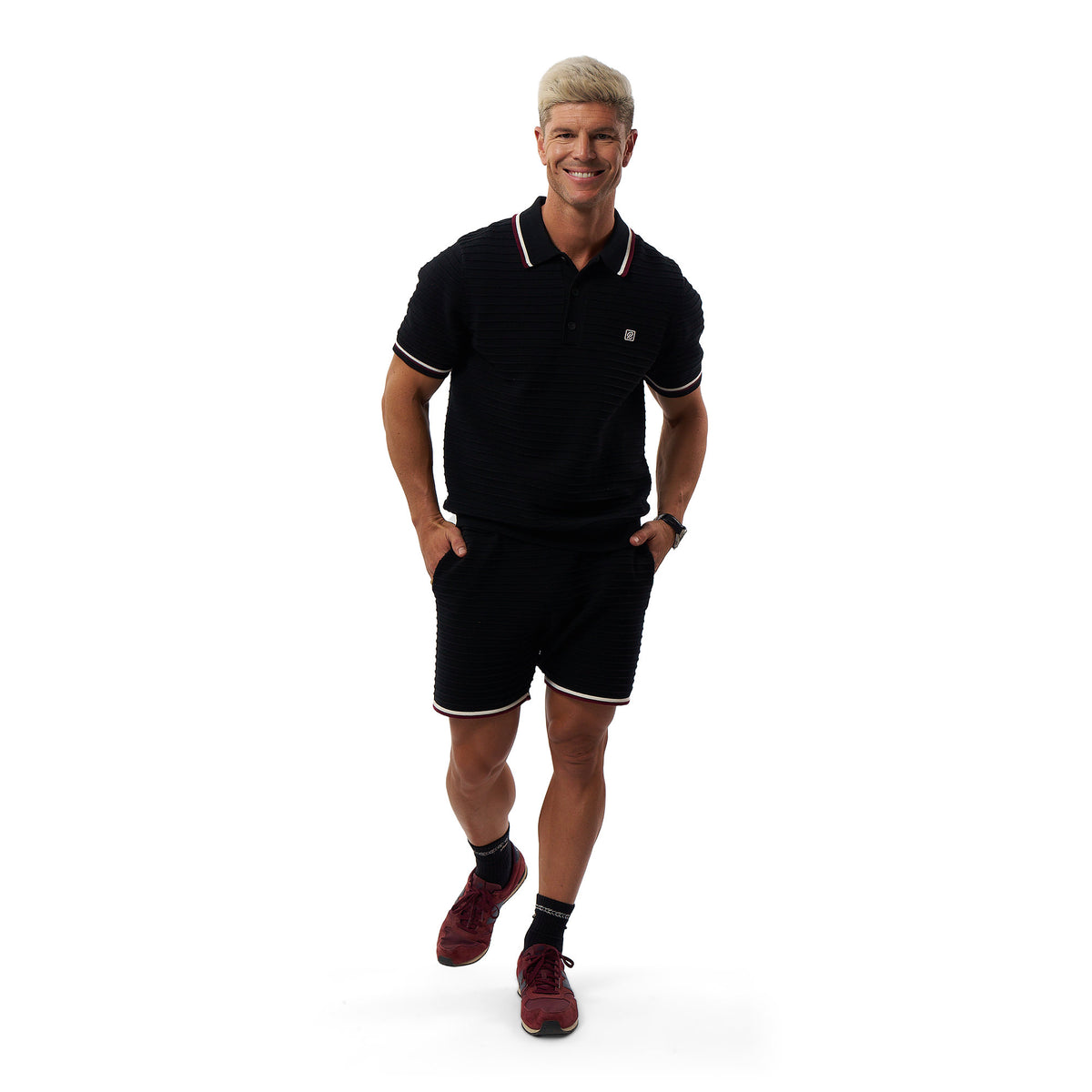 A man modeling a black short-sleeve polo shirt and matching black shorts with white trim, paired with dark maroon sneakers, against a plain white background.