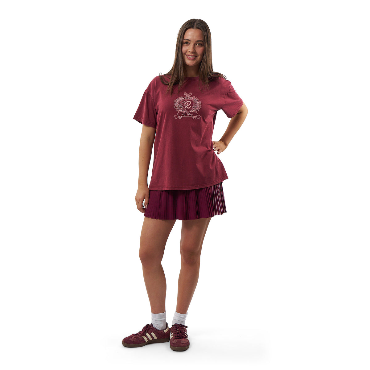 A young woman modeling a matching burgundy athletic set consisting of a short-sleeve t-shirt with a crest graphic and a pleated mini skirt, paired with burgundy sneakers and white socks, on a white background.