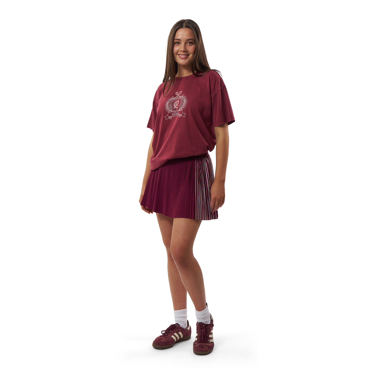 A young woman modeling a matching burgundy athletic set consisting of a short-sleeve graphic t-shirt and a pleated mini skirt, paired with burgundy sneakers, white socks, and standing against a plain white background.