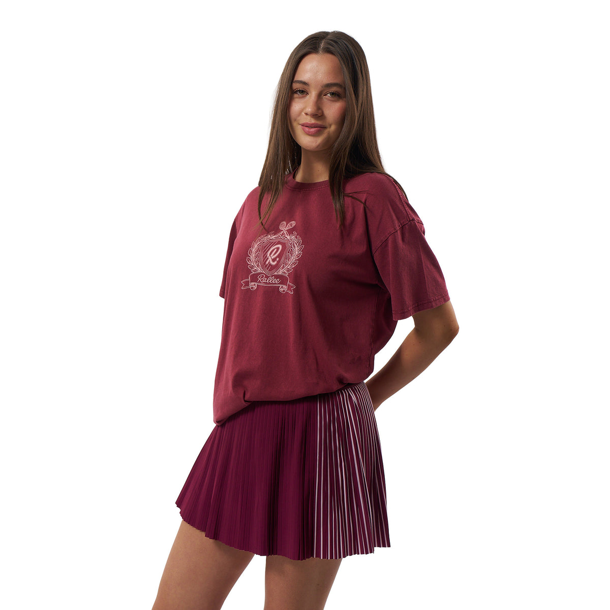 A young woman modeling a maroon short-sleeve t-shirt with a white crest graphic on the chest, paired with a matching pleated maroon mini skirt, against a plain white background.
