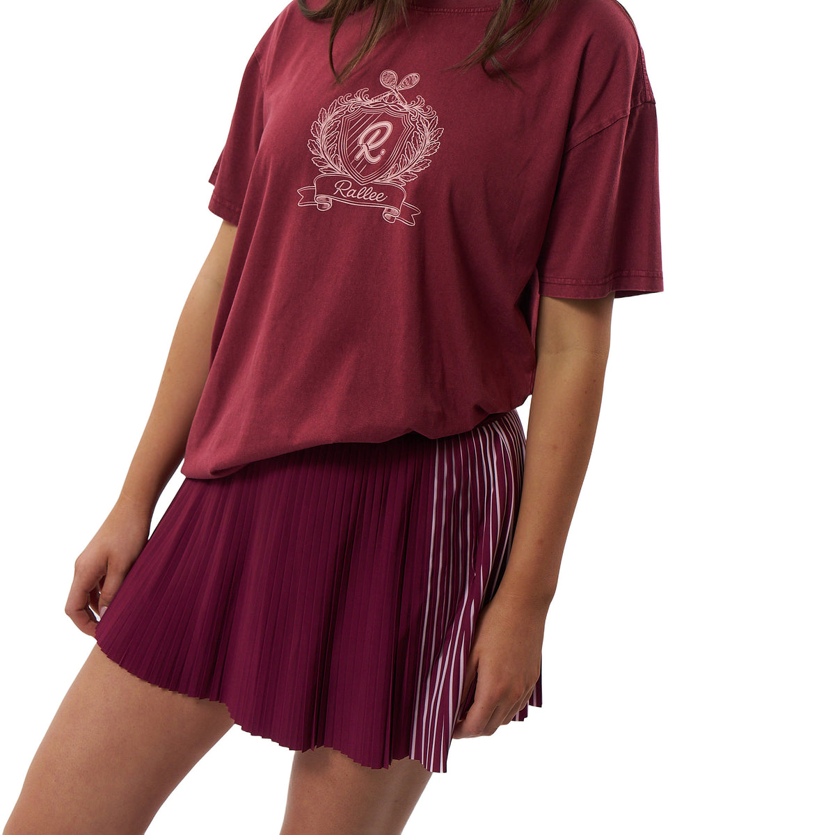 A model wearing a maroon short-sleeve graphic t-shirt with a white crest and the word 'Rallee' on the chest, paired with a matching pleated maroon skirt with white side stripes, against a plain white background.