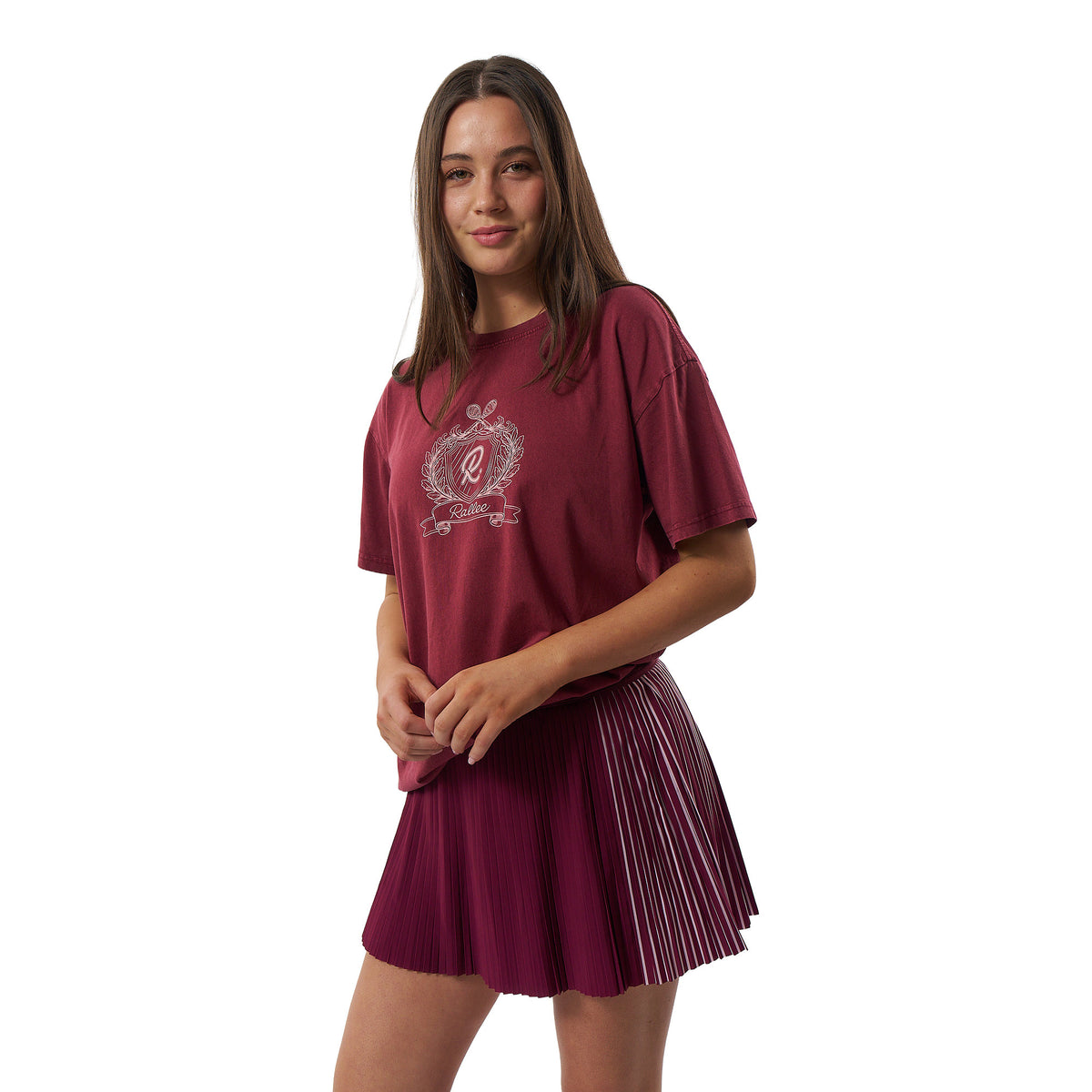 A young woman modeling a short-sleeve t-shirt and pleated mini skirt set in a deep maroon color, featuring a white crest graphic on the chest, set against a plain white background.