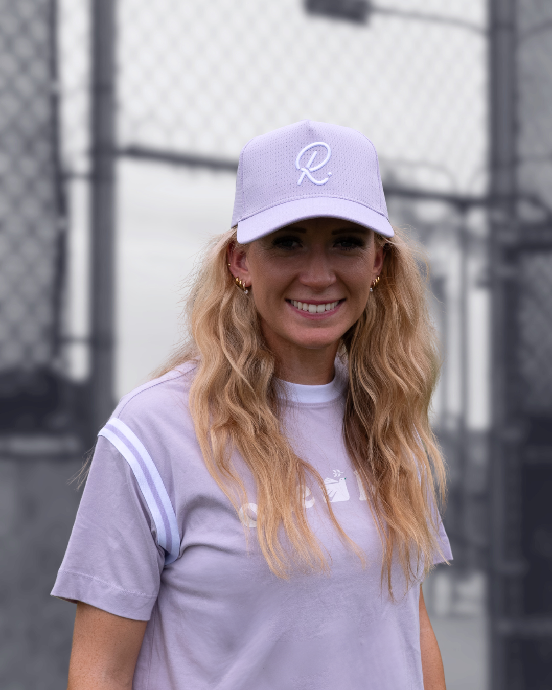 Woman wearing a light purple cap and t-shirt with a logo, standing in front of a tennis court