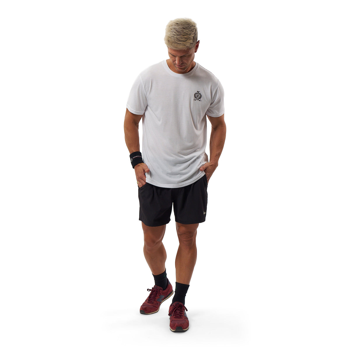 A man wearing a white short-sleeve athletic t-shirt with a small black chest logo, black athletic shorts, black socks, and red running shoes, standing against a solid white background.