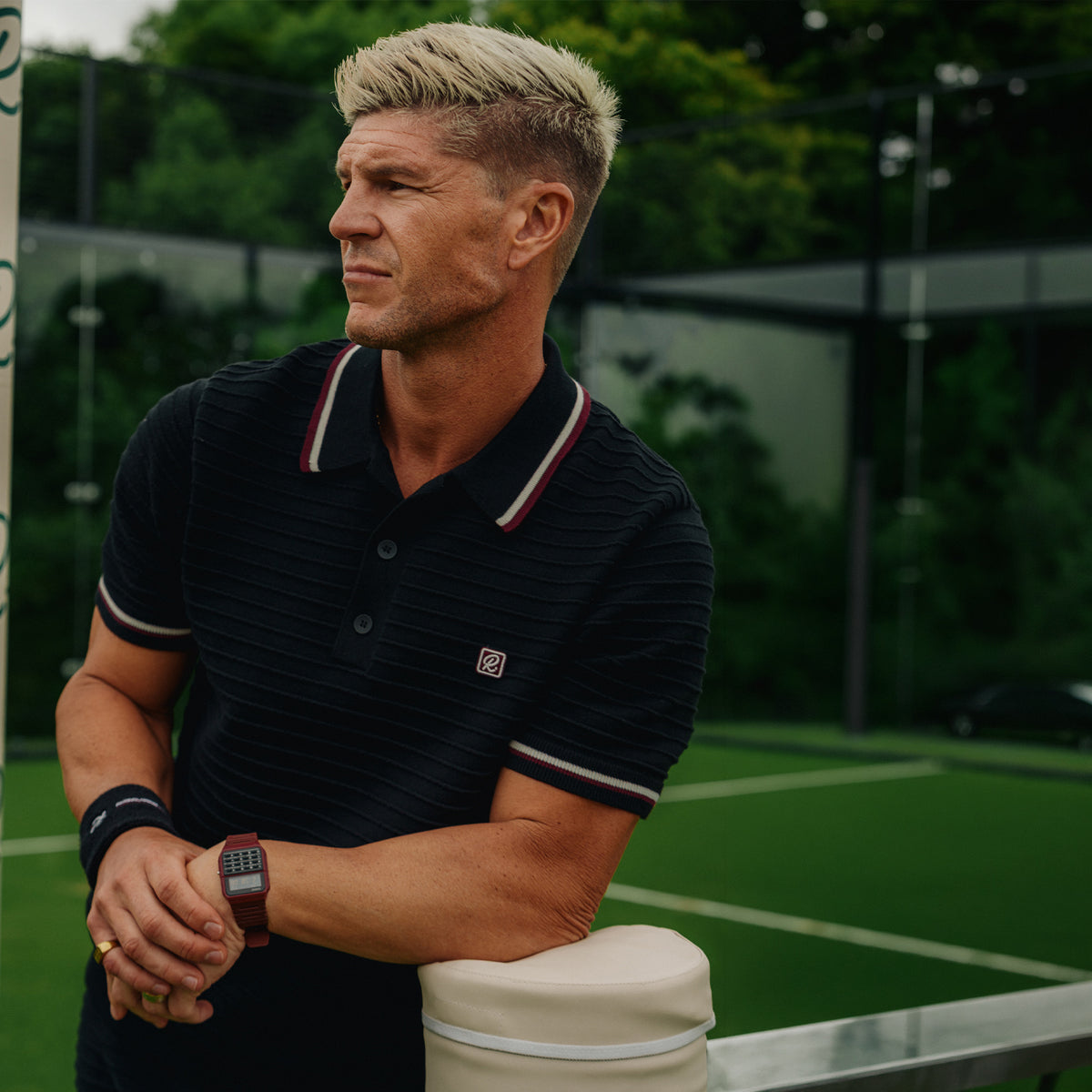 A man wearing a dark, ribbed polo shirt with white and red striped collar and sleeve trim, leaning on a white tennis racket on an outdoor tennis court.
