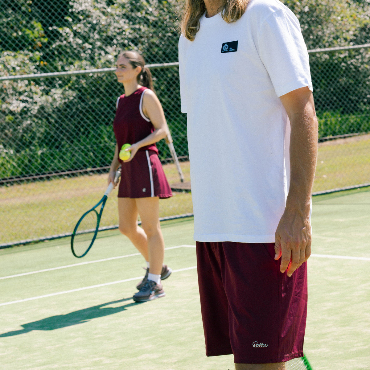 A man in a white t-shirt and maroon athletic shorts stands on a green tennis court, with a woman in a matching maroon tennis outfit holding a racket in the background near a chain-link fence.