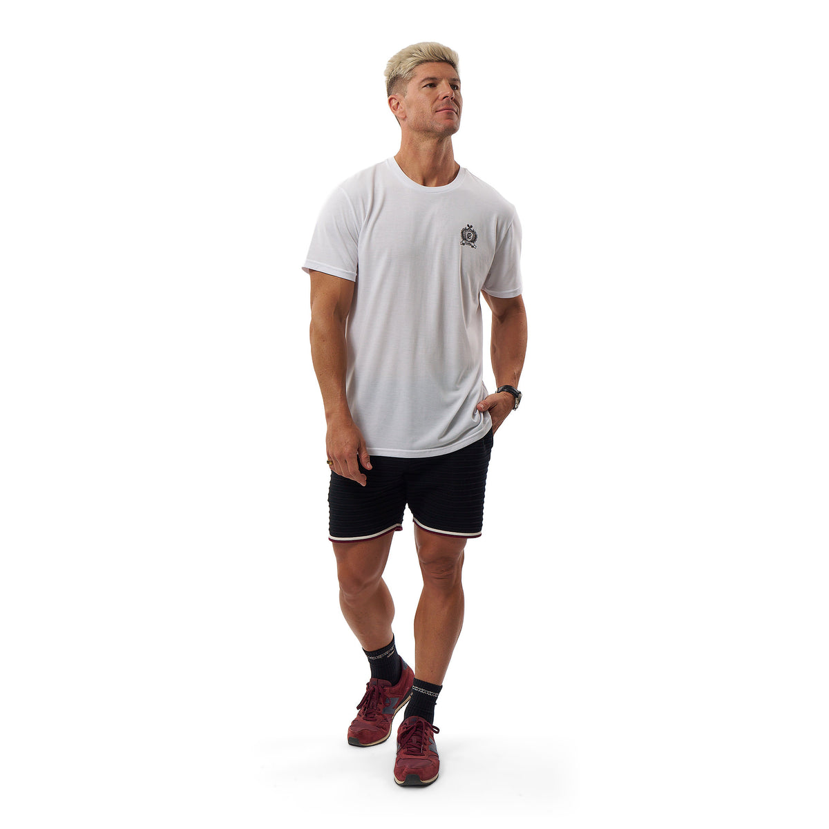 A man wearing a white short-sleeve athletic t-shirt with a small chest logo, black athletic shorts with white trim, dark socks, and maroon running shoes, standing against a solid white background.