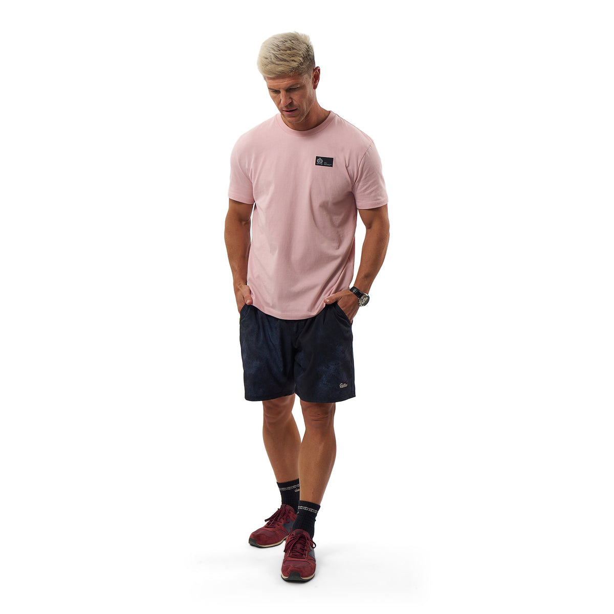 A man modeling a light pink crewneck t-shirt with a small black chest logo, paired with dark navy athletic shorts, dark socks, and burgundy athletic shoes, against a solid white background.