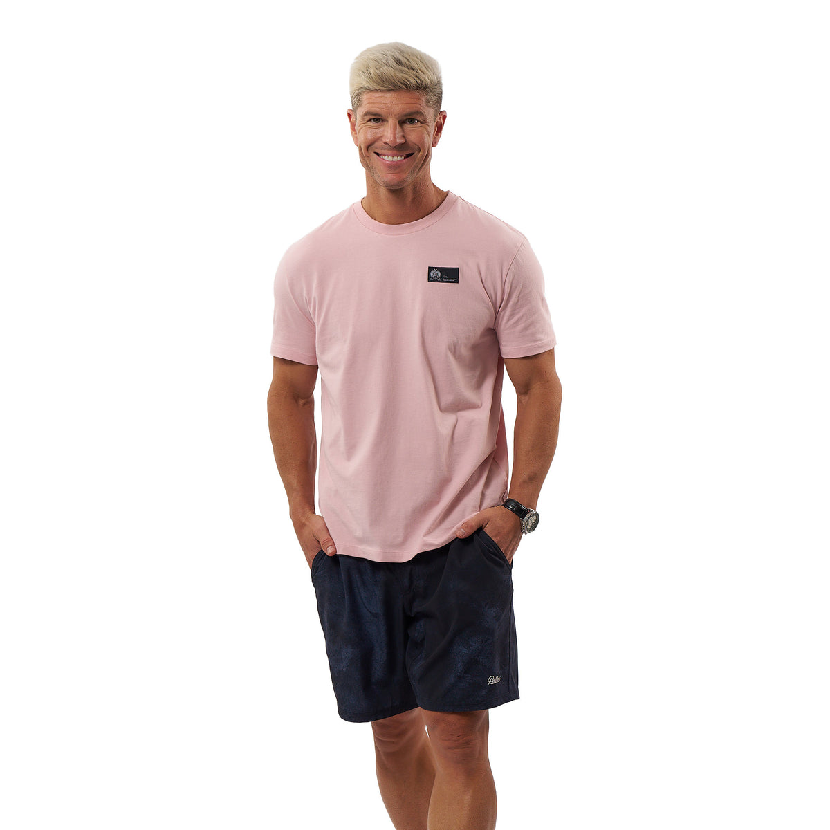 A man wearing a light pink crew-neck t-shirt with a small black chest logo and dark navy patterned shorts, standing against a solid white background.