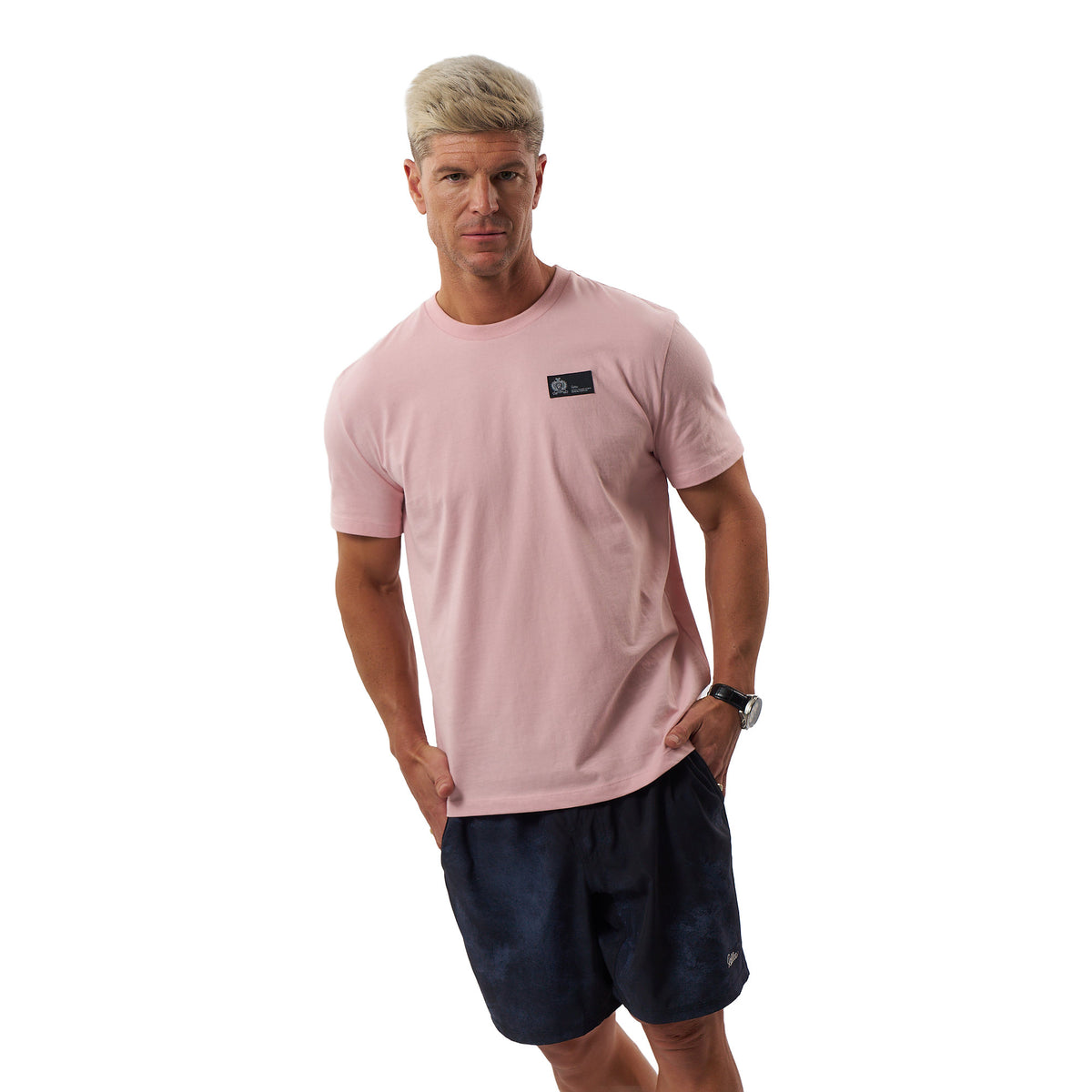 A man wearing a light pink crew-neck t-shirt with a small black rectangular logo patch on the left chest, paired with dark navy athletic shorts, photographed against a solid white background.