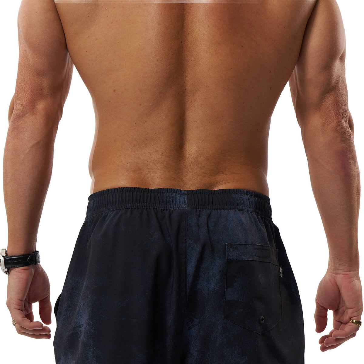 Rear view of a shirtless man wearing dark navy athletic shorts with an elastic waistband and a back pocket, against a plain white background.