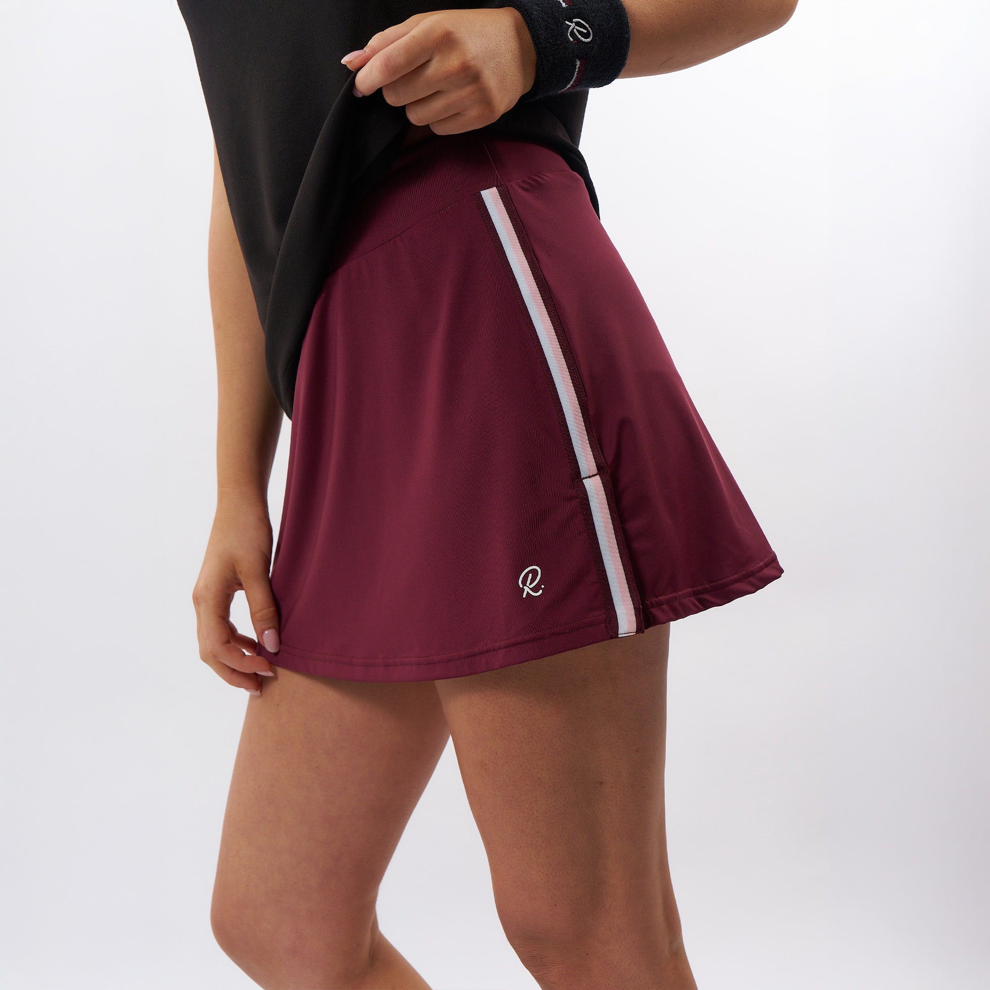 A close-up of a model wearing a maroon athletic tennis skirt with a white vertical stripe and a white embroidered logo, paired with a black top.