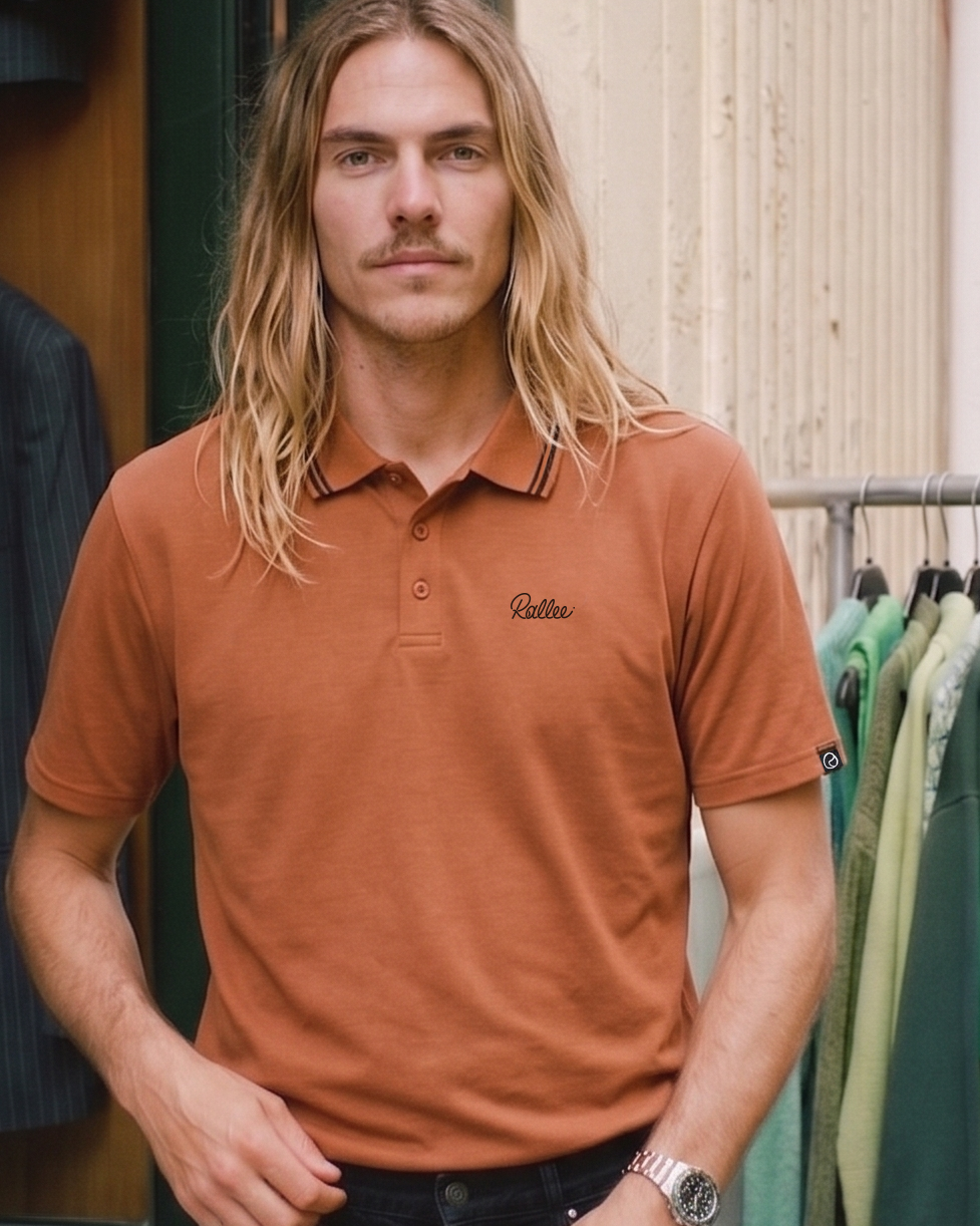 Man wearing a clay polo shirt with  Rallee logo, standing in front of a clothing rack.