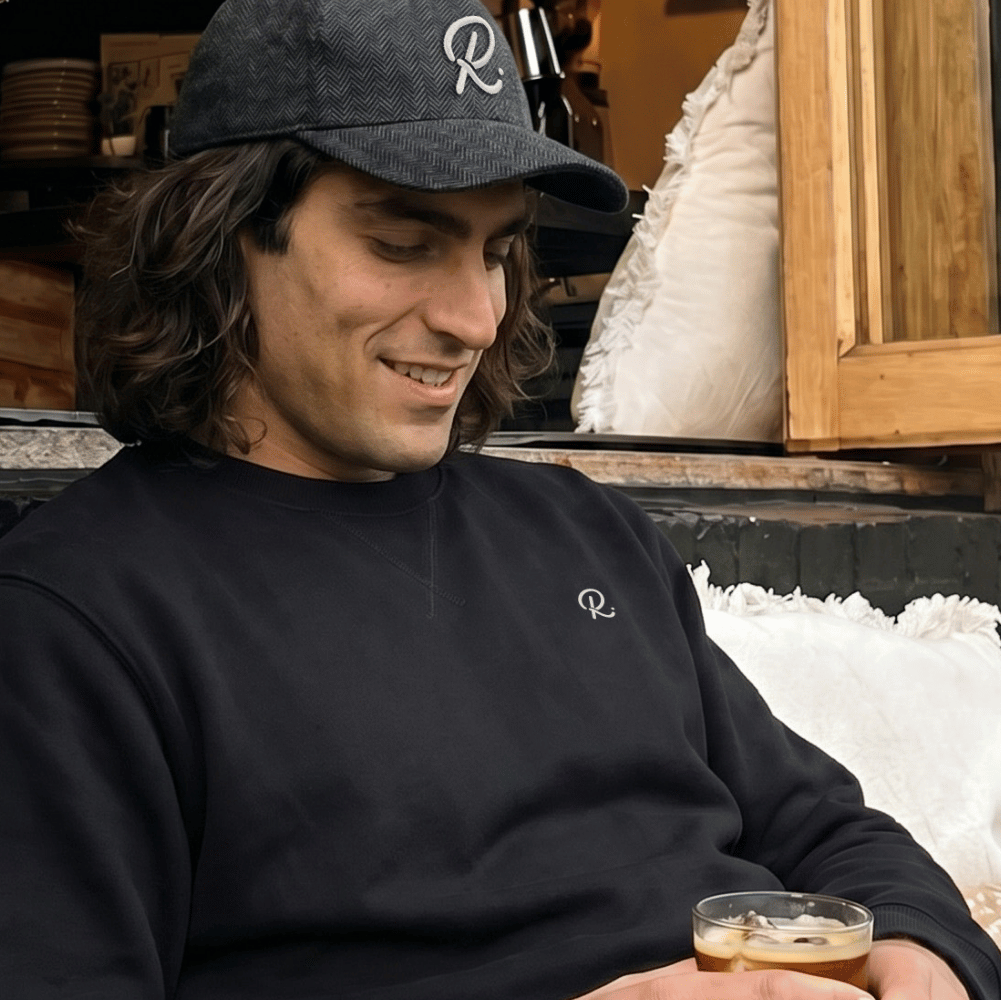 Man wearing a black cap and sweatshirt, holding a drink, sitting on a couch.