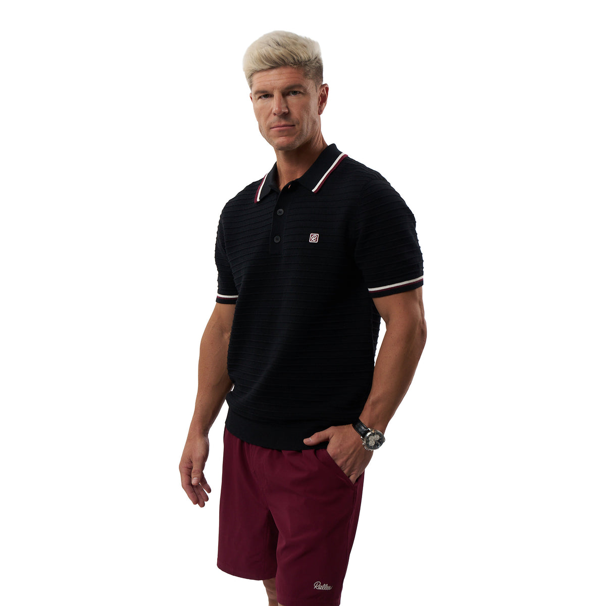 A man modeling a black short-sleeve polo shirt with white and red striped trim on the collar and sleeve cuffs, paired with burgundy athletic shorts, against a plain white background.