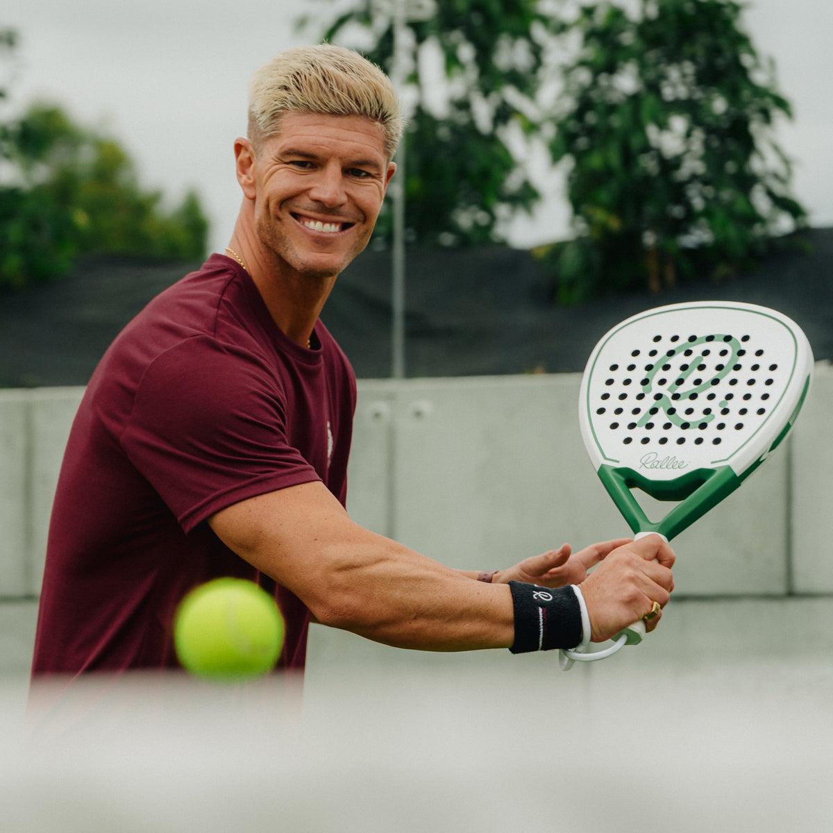 A smiling blonde man in a maroon t-shirt holds a green and white padel racket with a tennis ball in the foreground, set against an outdoor court background.