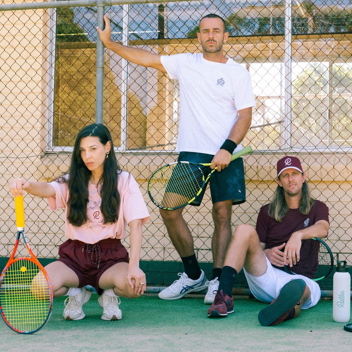 Three people on a tennis court holding tennis rackets, posing in front of a chain-link fence. The person on the left is crouching in a pink t-shirt and maroon shorts, holding an orange and yellow racket.