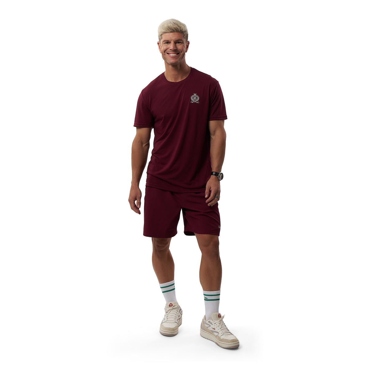 A man modeling a matching burgundy athletic set consisting of a short-sleeve t-shirt and shorts, paired with white socks and sneakers, against a plain white background.