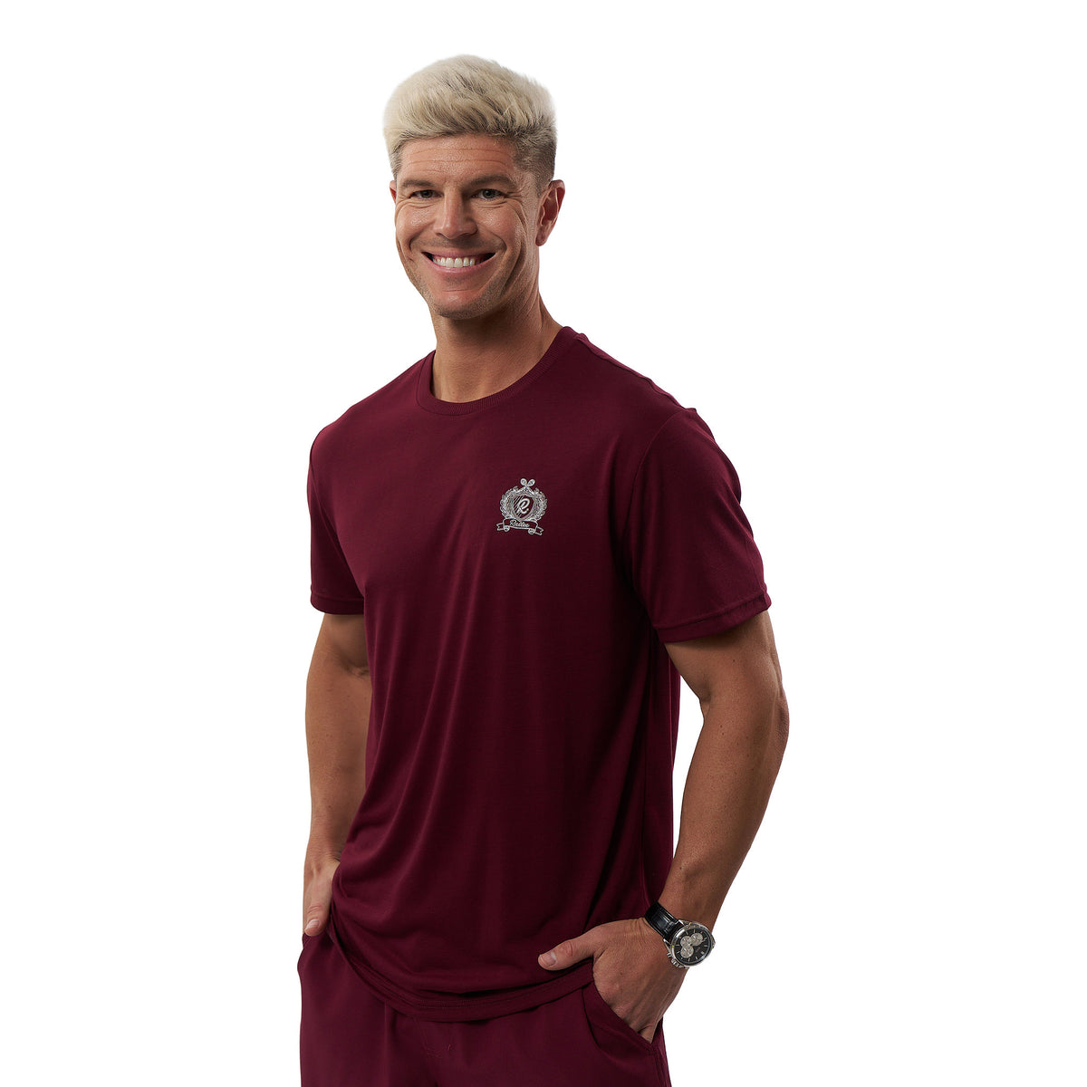A smiling man with short blond hair wearing a maroon short-sleeve athletic t-shirt with a small white crest logo on the left chest, paired with matching maroon shorts, against a plain white background.