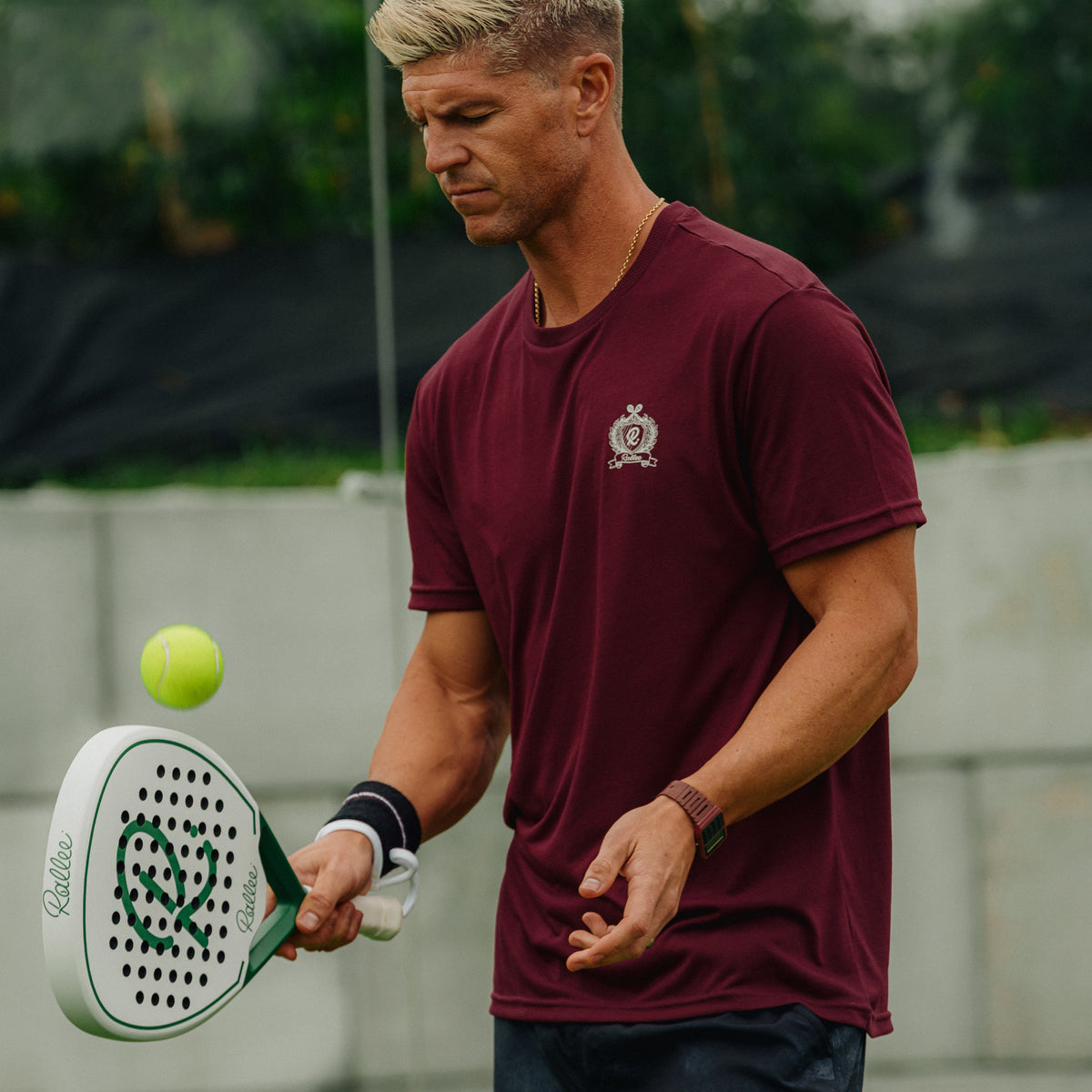 A man in a maroon short-sleeve athletic t-shirt with a small crest logo on the chest, holding a padel racket and preparing to hit a yellow tennis ball. The background shows a blurred outdoor court setting.