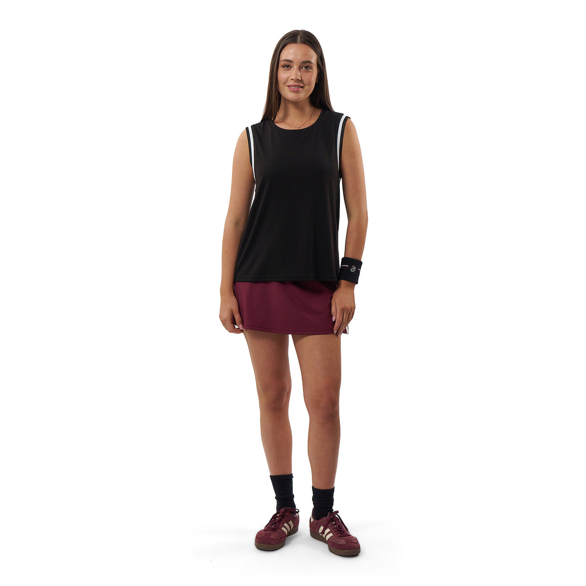 A woman modeling a sleeveless black athletic tank top with white shoulder stripes, paired with a burgundy athletic skirt, black socks, and burgundy running shoes against a plain white background.