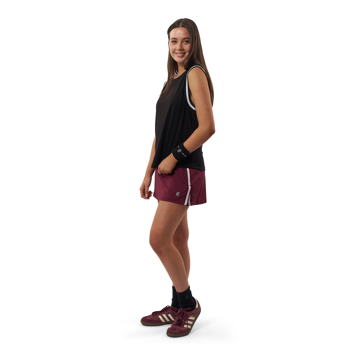 A young woman posing in athletic wear, featuring a black sleeveless tank top with white trim, maroon athletic shorts with white stripes, maroon Adidas sneakers, and black socks, set against a plain white background.