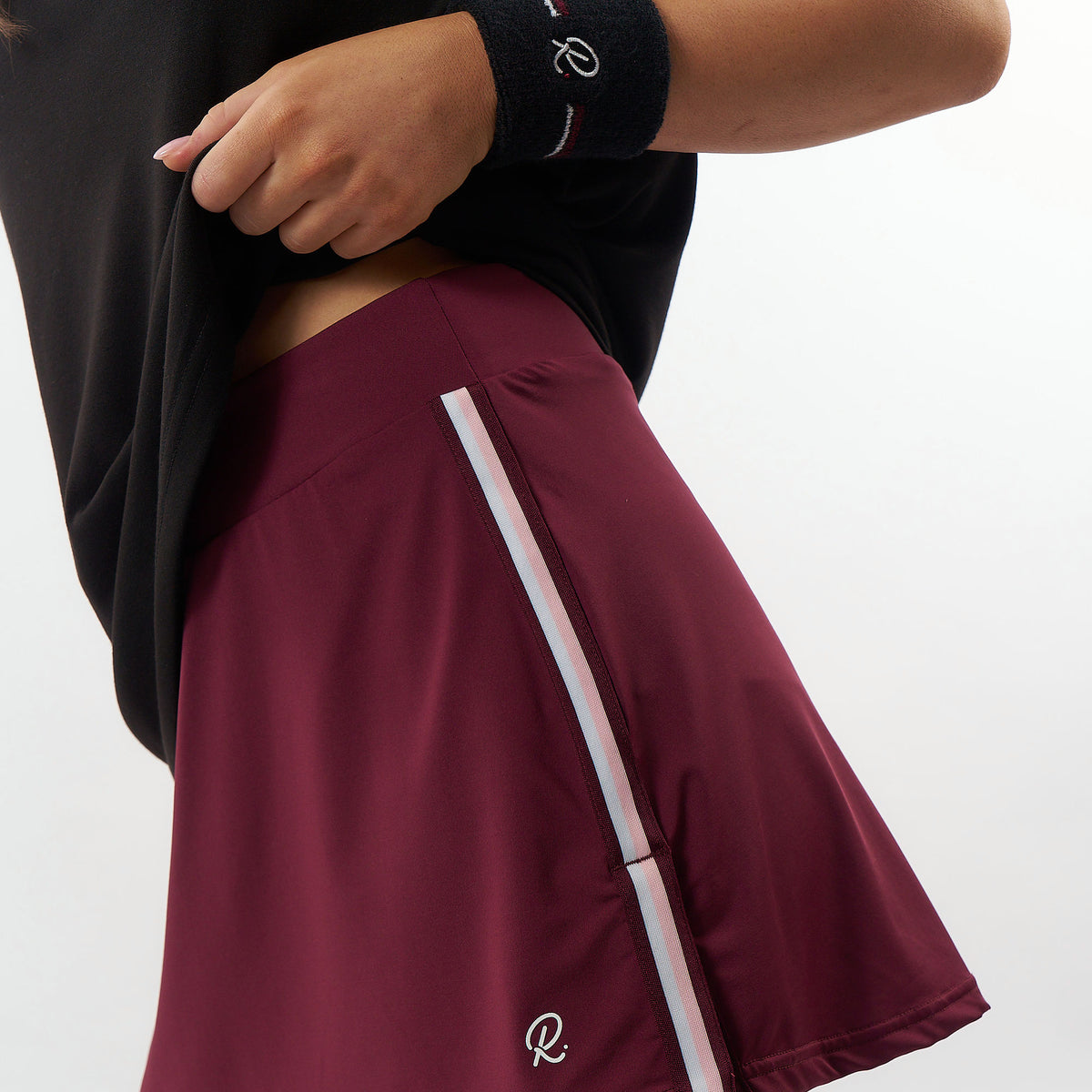 A close-up of a person wearing a maroon athletic skirt with a white side stripe and a white embroidered logo, paired with a black top and black wristband.