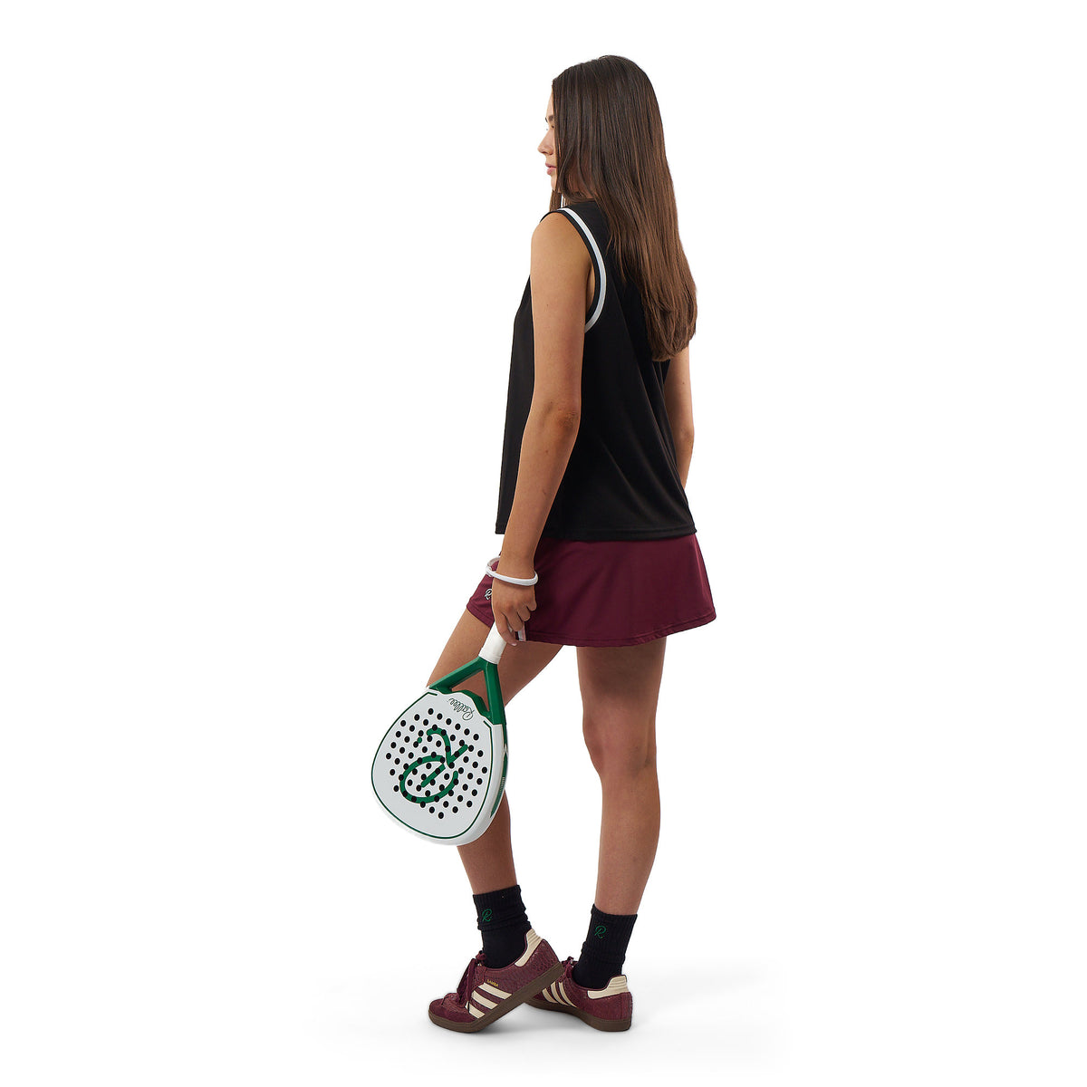 A woman in a black sleeveless top and burgundy tennis skirt stands in profile holding a white and green padel racket, photographed against a clean white background.