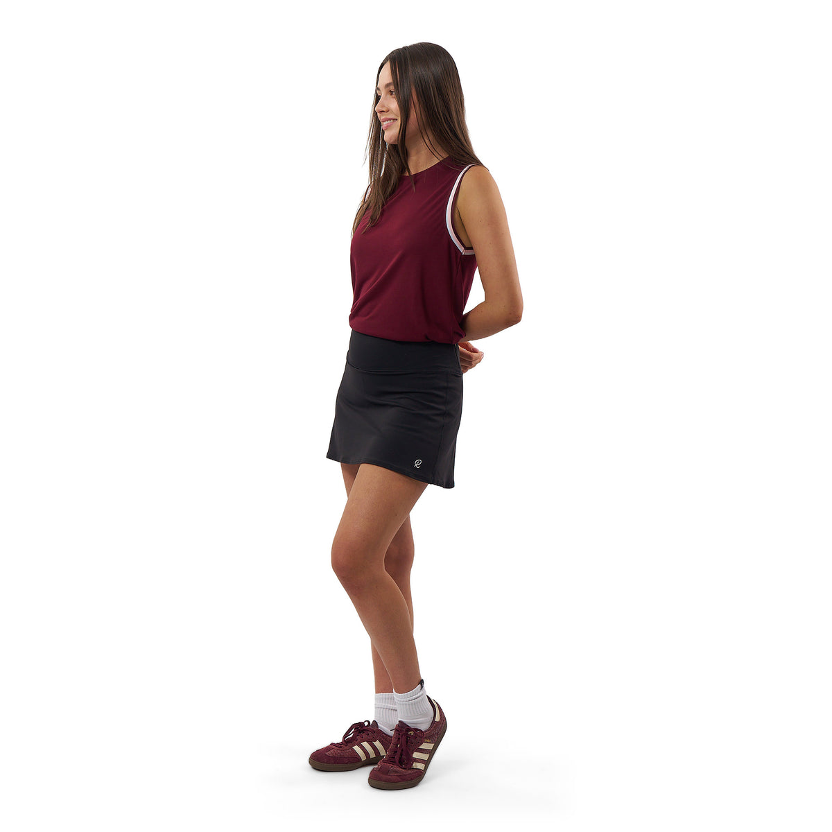A woman modeling an athletic sleeveless top in burgundy with white trim, paired with a black tennis skirt and burgundy running shoes, shown from a side profile against a white background.