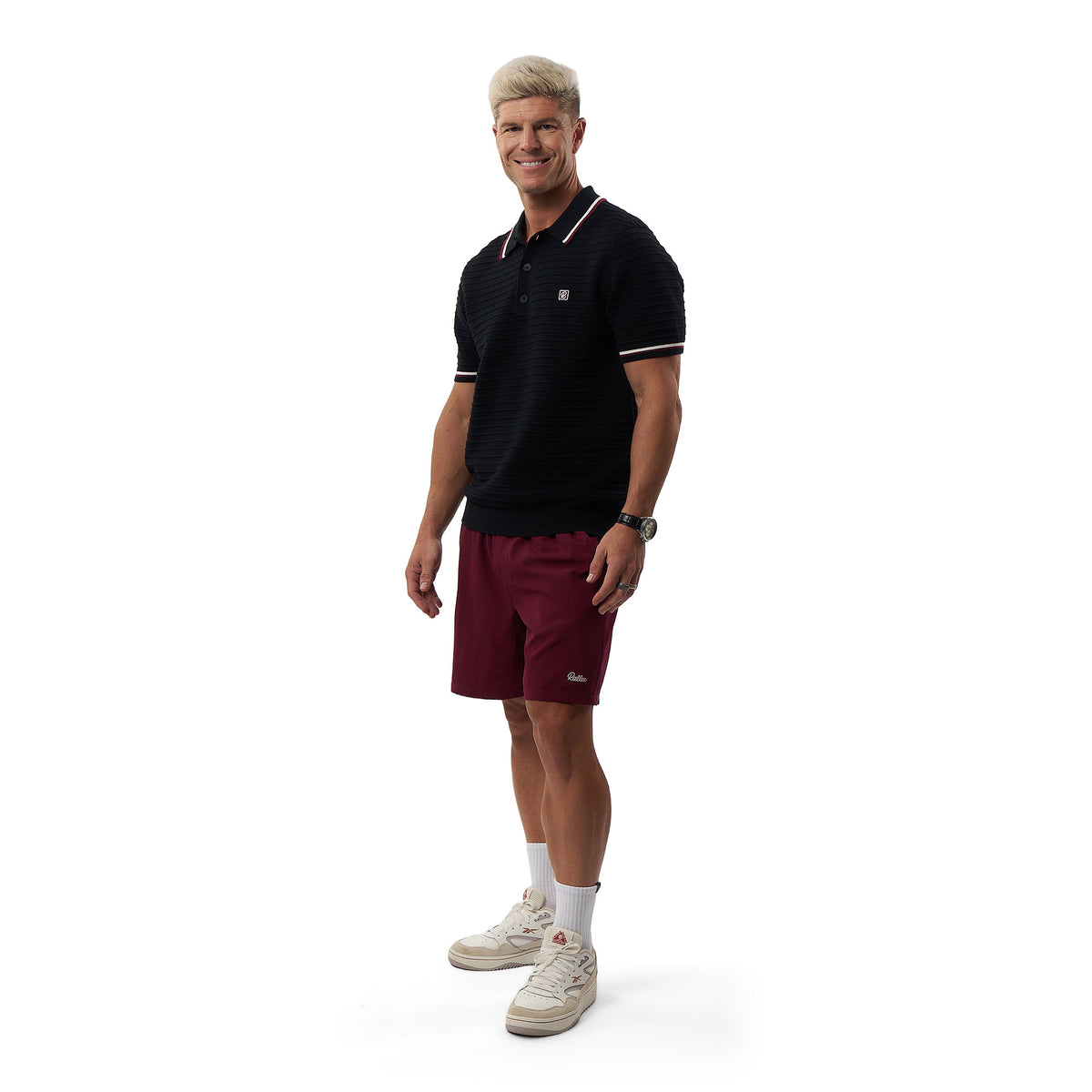 A man modeling a black short-sleeve polo shirt with white trim, maroon athletic shorts, white socks, and beige sneakers against a plain white background.