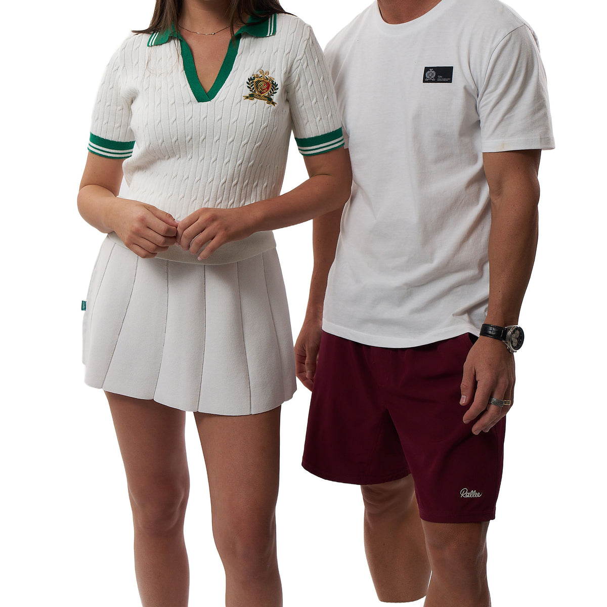 A man and a woman modeling white athletic outfits from Gallele. The woman wears a white textured short-sleeve polo shirt with green trim and a matching pleated white skirt.