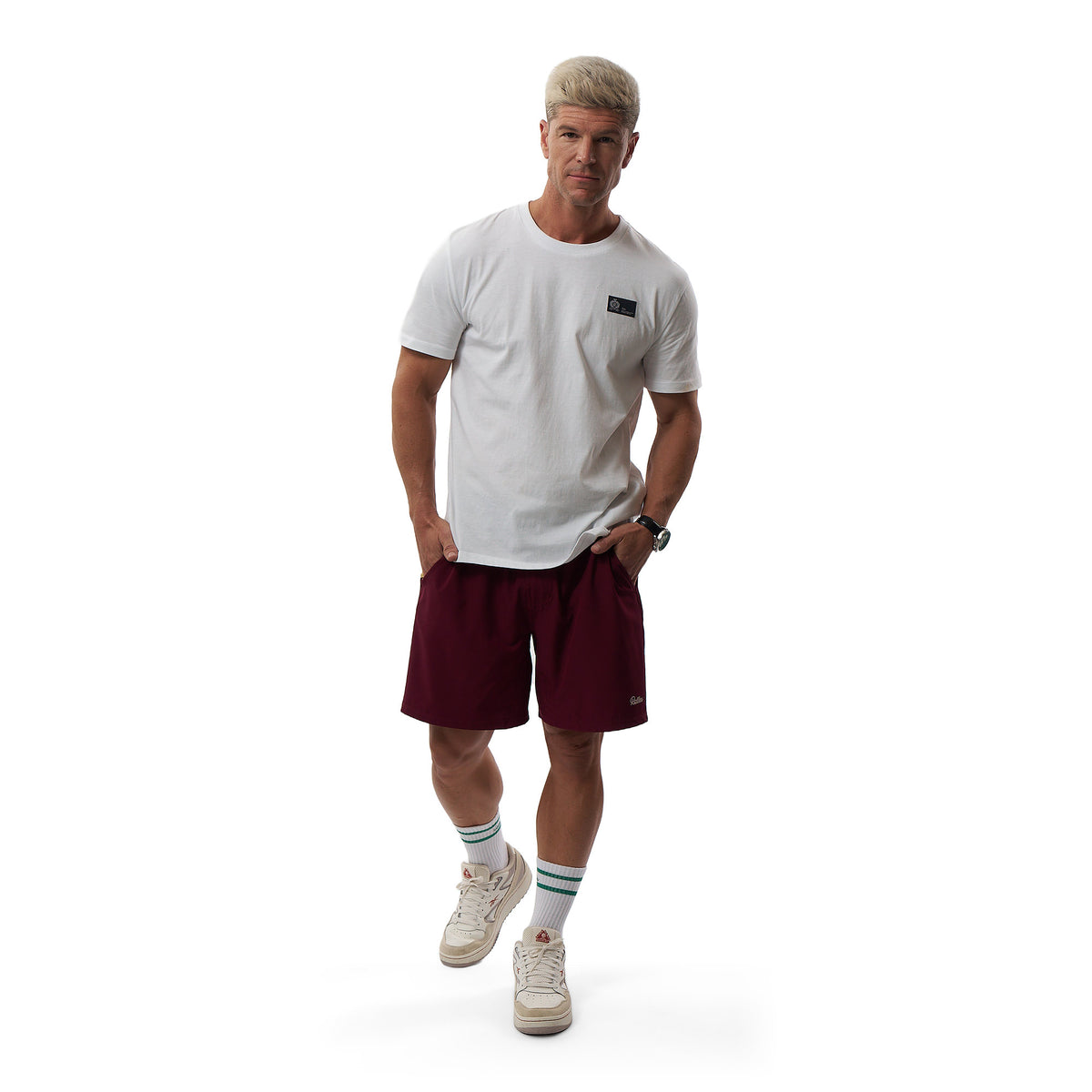 A man modeling a white crewneck t-shirt with a small black chest logo, paired with burgundy athletic shorts, white sneakers, and striped socks, against a solid white background.