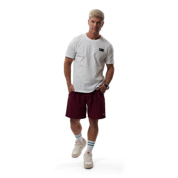 A man modeling a white crewneck t-shirt with a small black chest logo, paired with burgundy athletic shorts, white sneakers, and striped socks, against a solid white background.