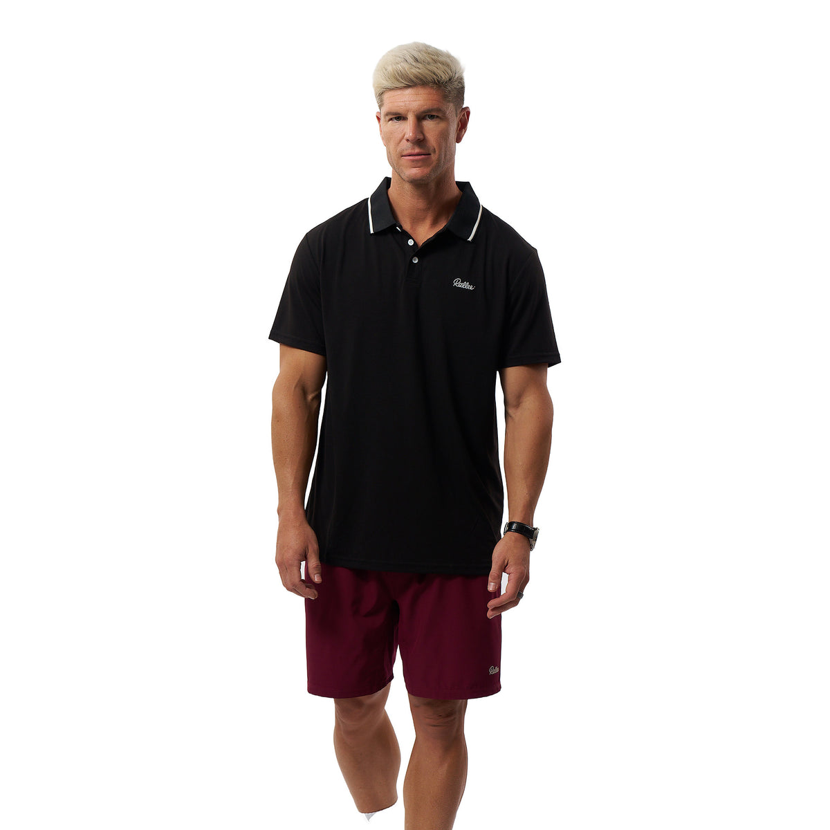 A man modeling a black short-sleeve polo shirt with white piping on the collar and a small white embroidered logo on the chest, paired with burgundy shorts, against a plain white background.