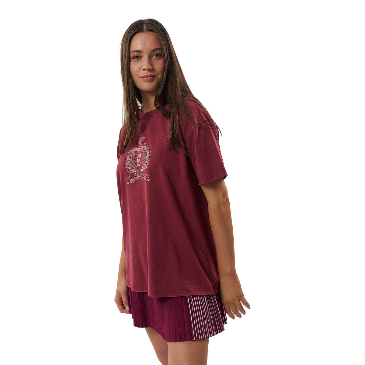 A woman modeling a short-sleeve crewneck t-shirt in a washed burgundy color, featuring a white graphic print on the left chest. She is also wearing a matching pleated skirt, set against a plain white background.