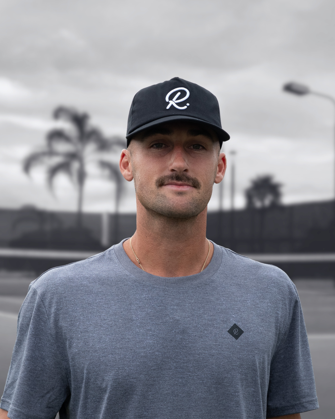 Man wearing a black cap with a white 'R' logo and grey t-shirt against a tennis court and palm trees background. 
