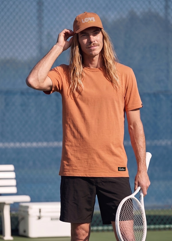A man with long blonde hair stands on a tennis court wearing an orange crewneck t-shirt, black shorts, and an orange baseball cap with 'LOVE' written on it. He is holding a white tennis racket in front of a blue chain-link fence.