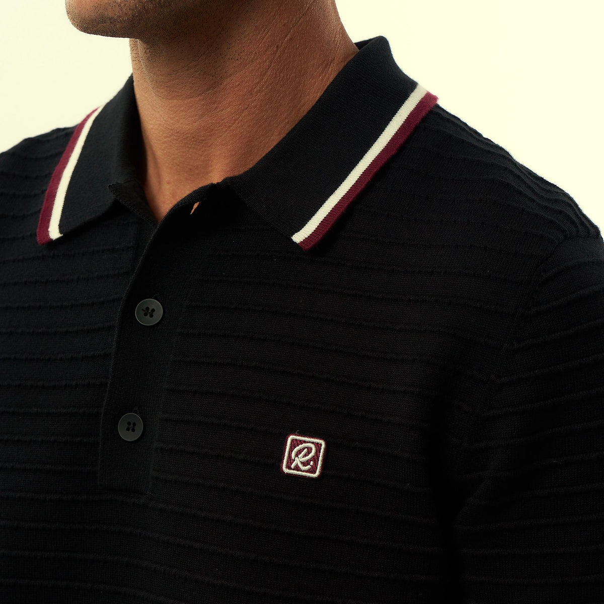Close-up of a man wearing a black ribbed polo shirt with a white and burgundy striped collar and a small embroidered logo on the chest.