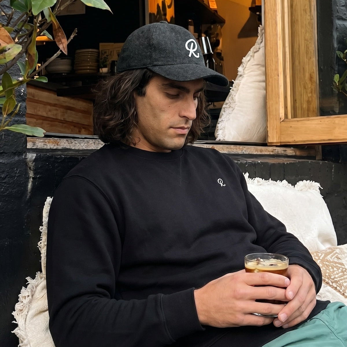 Man sitting outdoors holding a drink, wearing a black cap and sweater.