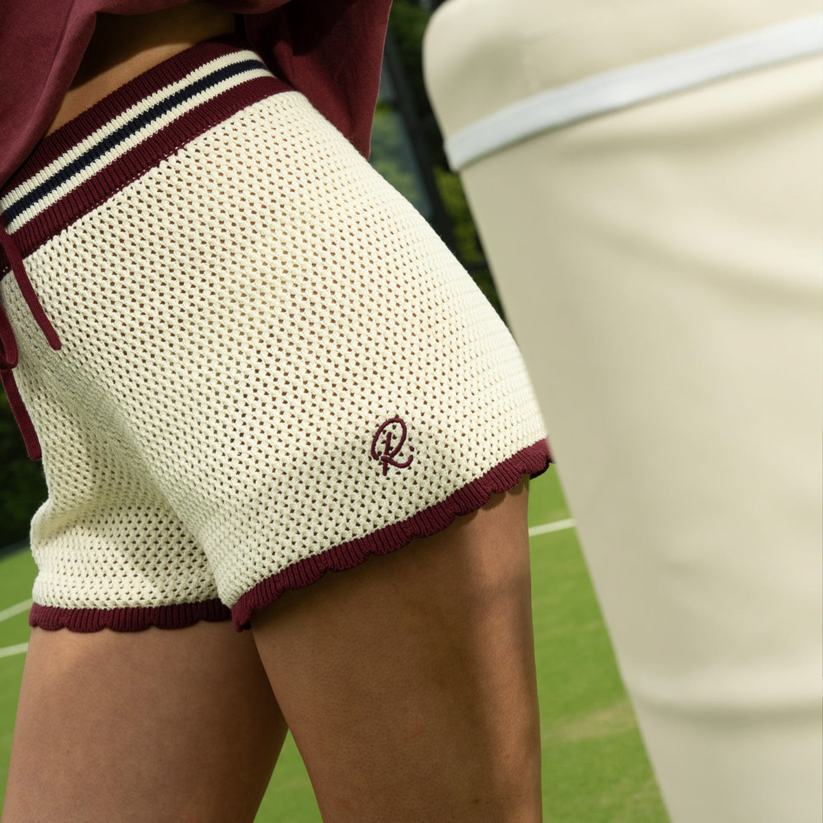 Close-up of a lady wearing cream knit shorts with burgundy trim and a small embroidered monogram logo, standing on a green tennis court.
