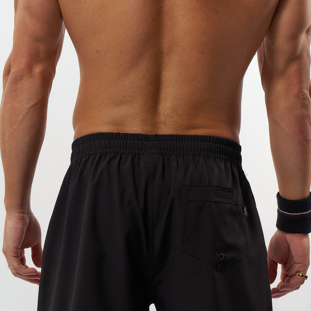 Rear view of a shirtless man wearing black athletic shorts with an elastic waistband and a side pocket, against a plain light background.